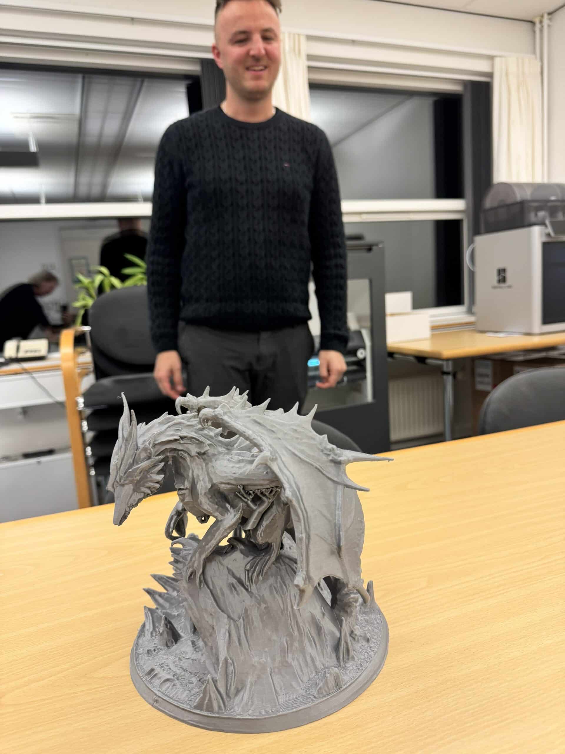 A detailed gray 3D-printed dragon sculpture stands on a wooden table in the foreground, while a smiling Edis stands behind it in an indoor workspace with chairs, windows, and equipment visible in the background.