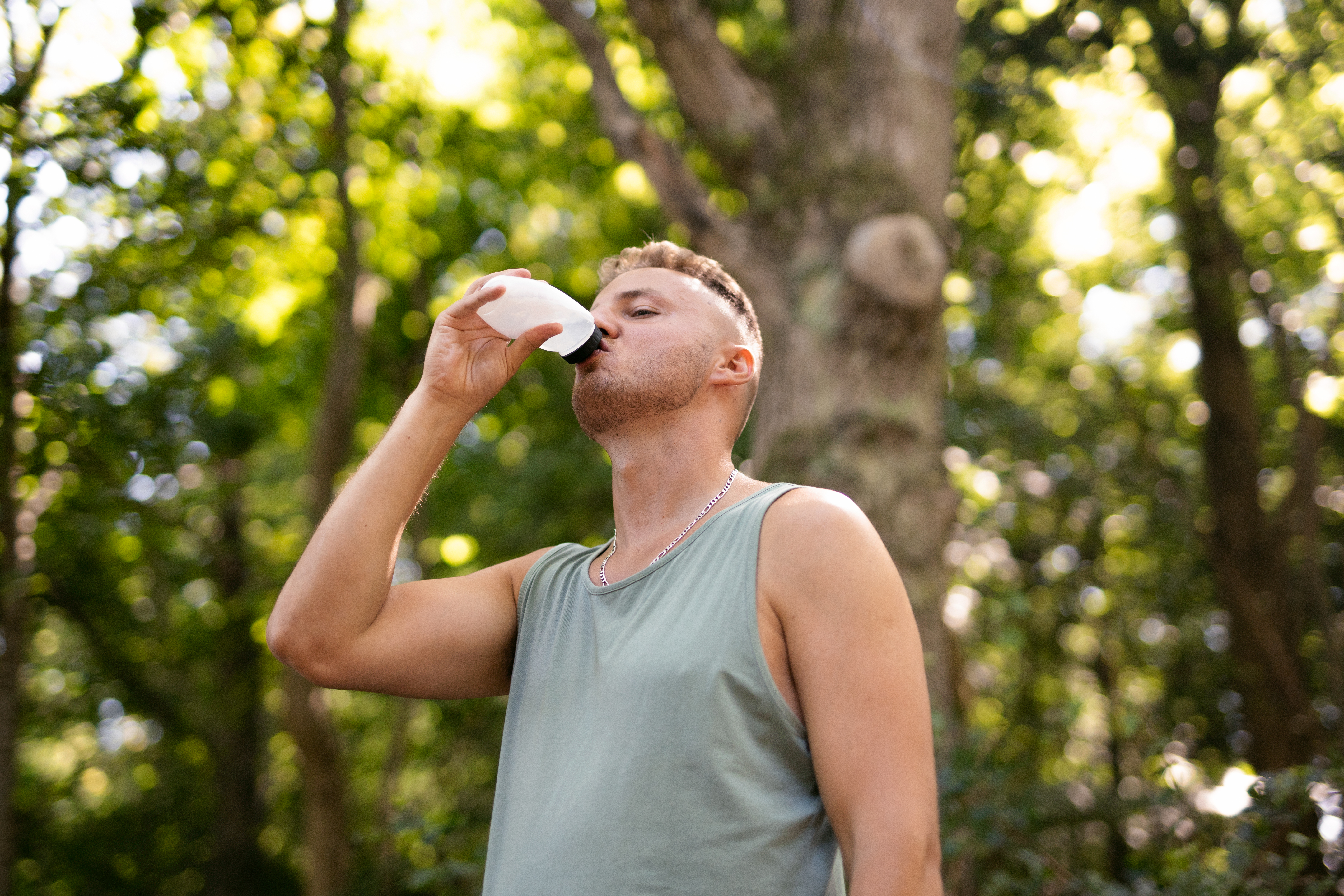 Edis in a sleeveless green top drinks from a white water bottle while standing in a sunlit forest, with trees and soft green foliage blurred in the background.
