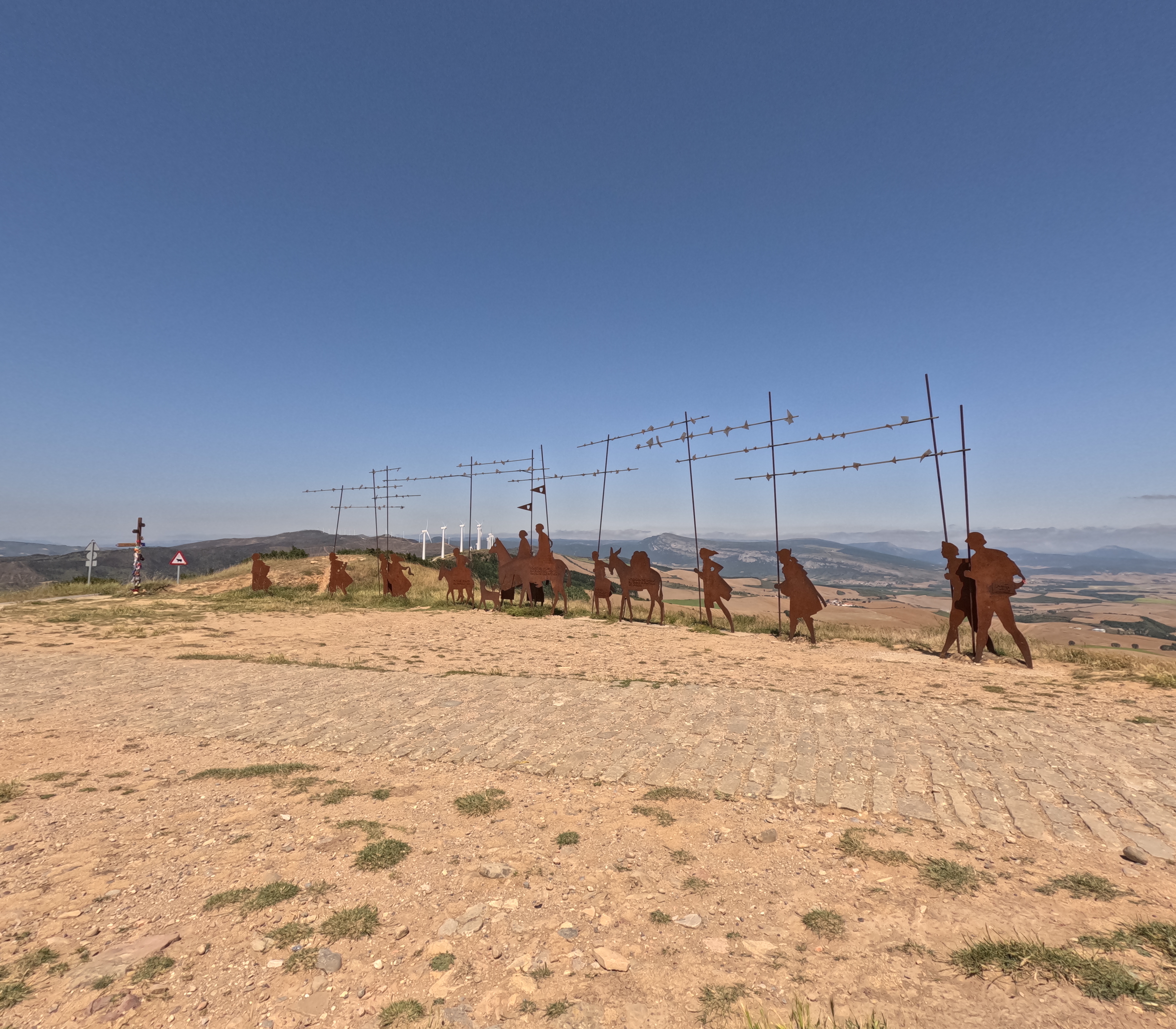 A line of rust-colored metal silhouettes depicting pilgrims with staffs stands along a hilltop path, set against a wide open landscape of fields and distant mountains under a clear blue sky.